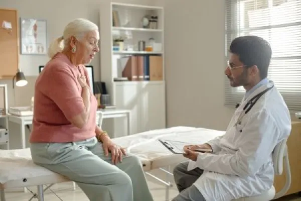 Senior woman consulting male doctor in clinic, describing chest pain during medical visit and receiving assessment.