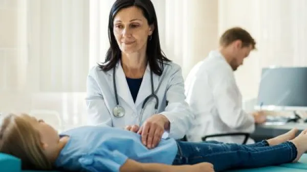 Female doctor in white coat palpating belly of girl lying on couch and talking to little patient