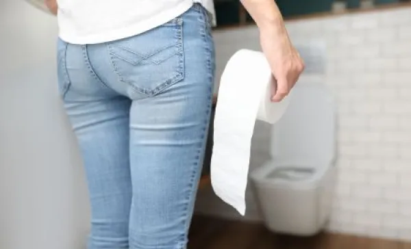 Woman in blue jeans stands with back and holds roll of toilet paper in her hand in front of toilet close-up.